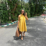 The Amalfi Halter Dress in golden ochre, walking with a tan tote bag